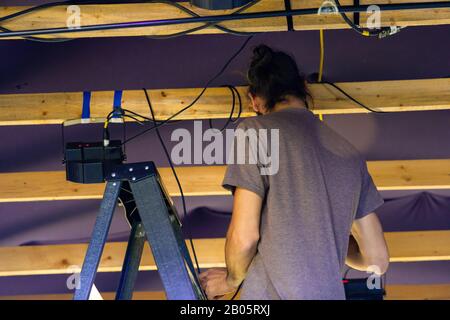 Una vista perdente e posteriore di un uomo al backstage di lavoro durante un festival multiculturale, lavoro a stagehand da scale che preparano attrezzature per musica set Foto Stock