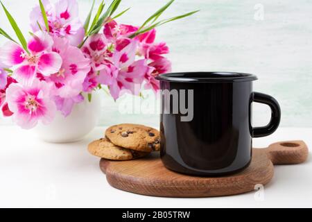 Mockup di tazza di caffè smalto nero campfire con biscotti e godezia rosa clarkia fiori. Tazza vuota per la promozione del design. Foto Stock