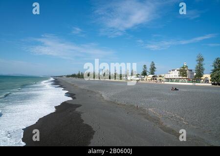 Napier Nuova Zelanda - 15 Febbraio 2020; Vista lungo la spiaggia grigia di pietra con passaggio pedonale, Marine Parade Gardens e soundshell con gli edifici della città in backgroun Foto Stock