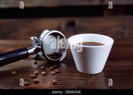 Una tazza di caffè nero, chicchi di caffè tostati e un supporto per macchina da caffè espresso su sfondo scuro. Foto Stock