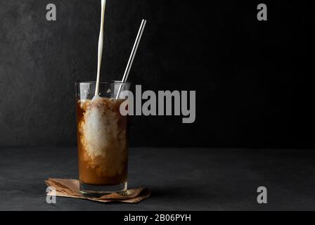 Caffè ghiacciato con panna da caffè in una tazza di vetro con una cannuccia metallica su sfondo scuro. Spazio di copia Foto Stock