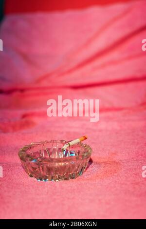 Posacenere con sigaretta bruciata sul lettino con asciugamano rosa. Foto Stock