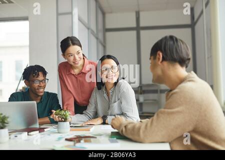 Team di giovani imprese che ascoltano il loro collega e sorridono che lavorano in team in ufficio Foto Stock
