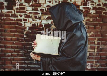 un stregone in un mantello nero e cappuccio contiene un libro tra le rovine Foto Stock