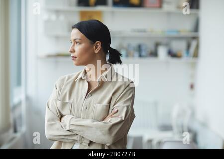 Ritratto di giovane donna d'affari pensosa in abiti casual in piedi in ufficio e guardare lontano Foto Stock