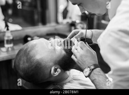 Utilizzo della macchina trimmer elettrica per lo styling di baffi e barba. Precisione e concentrazione del barbiere professionale. Processo di lavoro nel moderno salone di parrucchiere per uomini. Vista ravvicinata ritagliata. Foto Stock