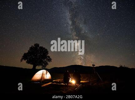 Estate notte campeggio, turismo e fotografia. Vista posteriore di due uomini seduti su grandi tronchi tra tenda turistica e macchina fotografica treppiede al fuoco ardente sotto cielo stellato scuro con costellazione di Milky Way. Foto Stock