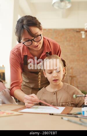 Giovane donna che tiene il pennello e aiuta la bambina a dipingere durante una lezione Foto Stock