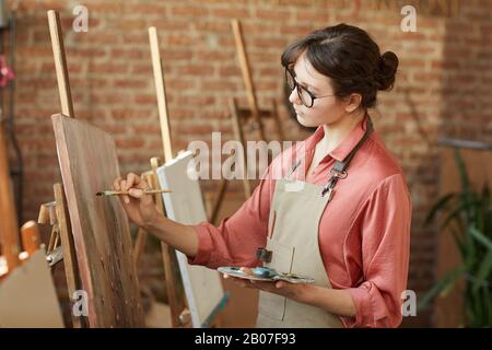 Giovane donna in occhiali pittura su cavalletto con vernici mentre si sta in piedi in studio d'arte Foto Stock