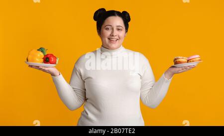 Sorrisi Plus Size Girl Holding Piatti Con Verdure Fresche E Ciambelle Foto Stock