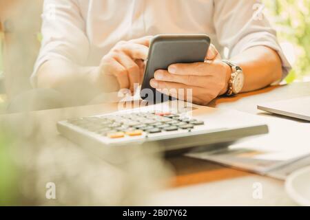 Le mani di un uomo d'affari che tiene lo smartphone con la calcolatrice su un tavolo di legno. Foto Stock