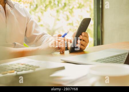 Le mani dell'uomo d'affari che tiene lo smartphone con il laptop sul tavolo di legno. Foto Stock