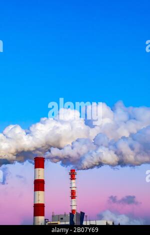 I camini della fabbrica inquinano l'aria, fumo spesso dipinto dal tramonto, il crepuscolo serale del cielo. Bellissimo paesaggio industriale verticale, ambientale prob Foto Stock