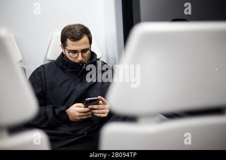 giovane adulto serio che guarda il suo smartphone in un posto metro. la fotocamera è nascosta tra due posti bianchi Foto Stock