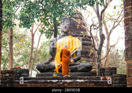 Un monaco buddhista in abito arancione prega davanti a una statua antica Foto Stock