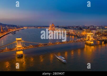 Budapest, Ungheria - veduta aerea del famoso Ponte delle catene Szechenyi illuminato a un'ora blu con barca turistica sul Danubio e il Parlamento a Foto Stock