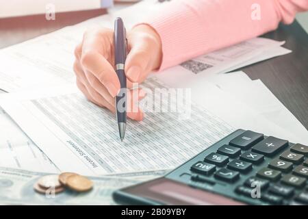 Closeup vista laterale sulla mano di contabile femminile che sta tenendo una penna e lavorando in un ufficio con una pila di documenti e tabelle. Calcolo di Foto Stock
