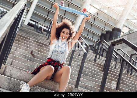 Giovane donna nella strada della città seduta su scale con Penny board guardando macchina fotografica confusa Foto Stock