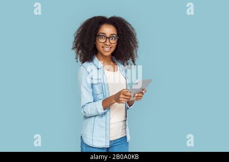 Stile Libero. Ragazza africana in occhiali in piedi isolato su Internet grigio navigazione su tablet digitale sorpreso Foto Stock