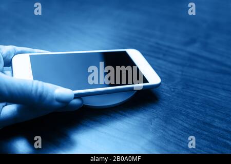 La mano tiene uno smartphone mobile per la ricarica in modalità wireless condividendo la batteria tramite il dispositivo in colore blu classico. Tecnologia di ricarica. Condivisione Della Carica Del Dispositivo Tecnico. Primo piano. Foto Stock