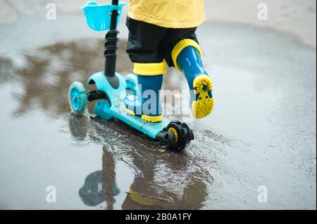 Un ragazzino felice in un impermeabile giallo guida uno scooter attraverso la pozza nel caldo giorno estivo dopo la pioggia. Tempo libero attivo per i bambini. Bambino che si diverte Foto Stock