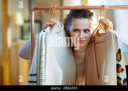 pensoso trendy 40 anni casalinga in maglione bianco e gonna a casa moderna nella soleggiata giornata invernale in buon umore scegliendo maglioni appesi su rame c. Foto Stock