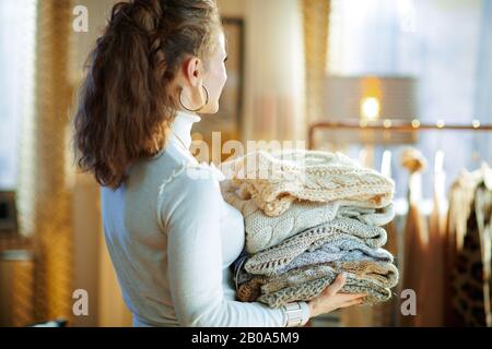 Vista da dietro elegante casa di mezza età in maglione bianco e gonna nella casa moderna nella soleggiata inverno giorno tenendo mucchio di maglioni. Foto Stock