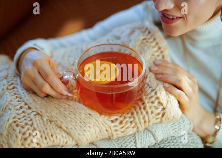 Vestirsi con la moderna donna di 40 anni in maglione bianco e gonna con pile di maglioni e tazza di tè al limone nel moderno soggiorno in vittoria soleggiata Foto Stock