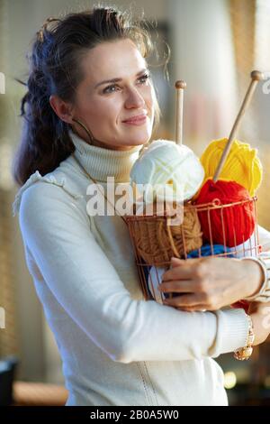 felice casalinga di 40 anni in maglione bianco e gonna con cesto con filo da maglia e aghi nel moderno soggiorno nella soleggiata giornata invernale. Foto Stock