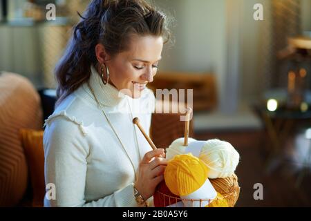 moderna casalinga sorridente di mezza età in maglione bianco e gonna con cesto con filo di maglia e aghi nella casa moderna nella soleggiata giornata invernale. Foto Stock