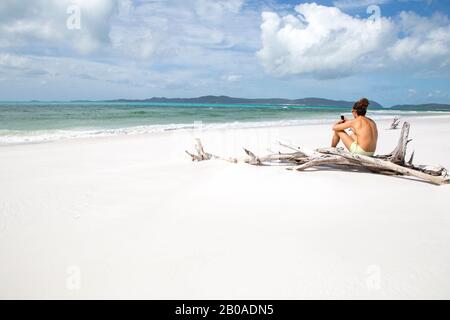 Uomo con smartphone, seduto sul log a spiaggia di sabbia bianca dell'Australia Foto Stock