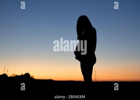 Una donna incinta tocca il ventre è silhoueted contro un sole colorato Foto Stock