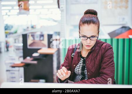 Giovane donna spara materiali di finitura in negozio di mobili. Indossa occhiali e giacca viola Foto Stock