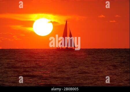 Una barca a vela naviga lungo le sagome dell'acqua contro un cielo di tramonto arancione in un tranquillo paesaggio naturale. Foto Stock