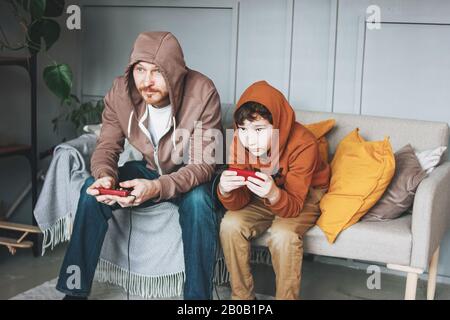 Padre e figlio Tween boy in hoodies giocare giochi per computer con joystick a casa tipica caucasica famiglia Foto Stock