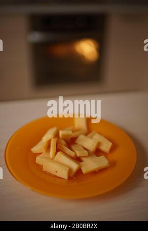 Pezzi di formaggio duro si trovano su un piatto sul tavolo da cucina. Forno di lavoro di fondo con luce calda. Foto Stock