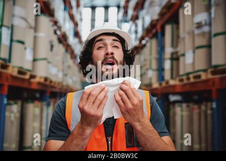 Giovane supervisore del magazzino caucasico starnutisce mentre si sente male di freddo Foto Stock