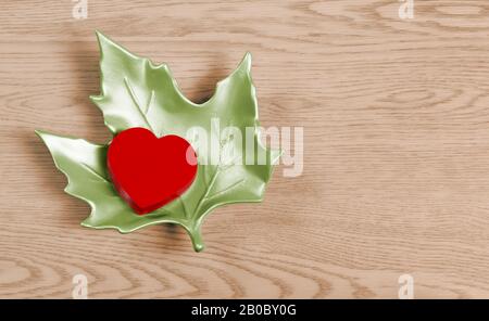 Scatola a forma di cuore rossa su una foglia di acero verde ceramica su sfondo di legno Foto Stock