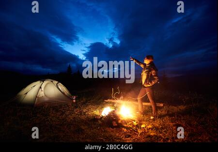 Donna attiva escursionista con un riposo di notte in campeggio in montagna, in piedi vicino al fuoco ardente e tenda turistica illuminata, che punta a sera cielo nuvoloso. Turismo, attività all'aperto concetto Foto Stock