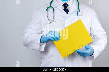 medico maschile in un cappotto bianco con bottoni, indossando guanti sterili blu, tenendo un notebook di carta gialla in mano su uno sfondo bianco studio Foto Stock