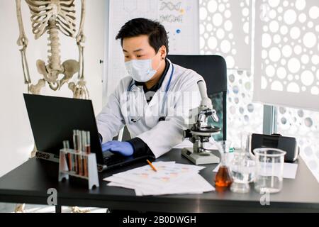 Lo scienziato asiatico giovane uomo in bianco cappotto, maschera e guanti fa biologia medicina o ricerca chimica, digitando su laptop, seduto al tavolo con Foto Stock