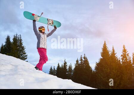 Ragazza sportiva è in piedi su una collina innevata che tiene il suo snowboard sopra la testa, foresta sullo sfondo, copia spazio, concetto di sport invernali estremi Foto Stock