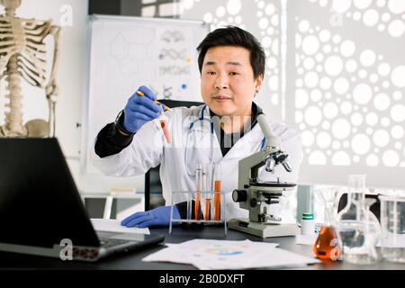 Medico coreano asiatico biochimico scienziato medico in camice bianco e guanti protettivi che tengono provetta con liquido rosso conduttore esperimento in Foto Stock