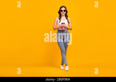 Foto a tutta lunghezza di attraente donna allegra buon umore a piedi strada bere caldo takeaway caffè indossare retro specifiche bianco tank-top jeans scarpe isolato Foto Stock