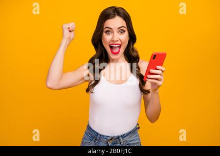 Foto di pazzo divertente signora tenere le mani del telefono sollevare pugno umore eccitato ha ottenuto molti gradises reposts commenti indossare bianco tank-top jeans isolato luminoso Foto Stock