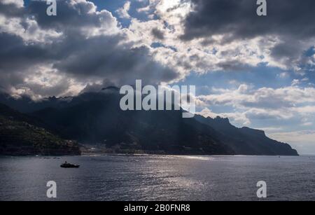 La luce del sole del mattino si infrange tra le nuvole, illuminando uno yacht sul mare calmo, le montagne della Costiera Amalfitana d'Italia Foto Stock