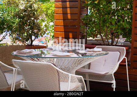 Tavolo con tovaglia bianca, posate e tovaglioli tessili sulla veranda esterna estiva del ristorante. Piante verdi in pentole sullo sfondo. Foto Stock