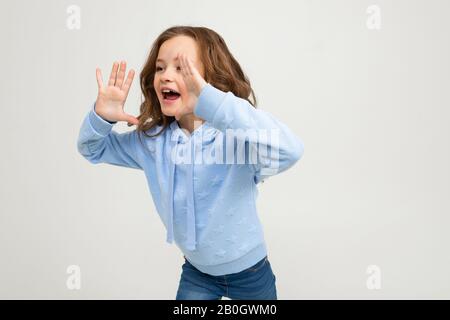 la ragazza adolescente europea in una felpa con cappuccio blu grida la notizia mentre tiene le mani in bocca su uno sfondo chiaro Foto Stock