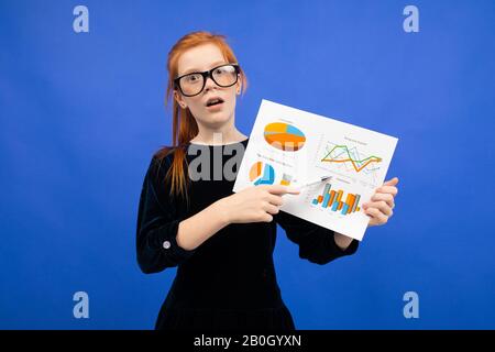 ragazza adolescente intelligente in occhiali e un abito nero mostra le statistiche con grafici a torta e grafici su uno sfondo blu Foto Stock