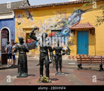 Sculture di eroi indipendenti in Plaza Trinidad di fronte all'arte di strada che decorano un edificio coloniale a Getsemani, Cartagena, Colombia Foto Stock